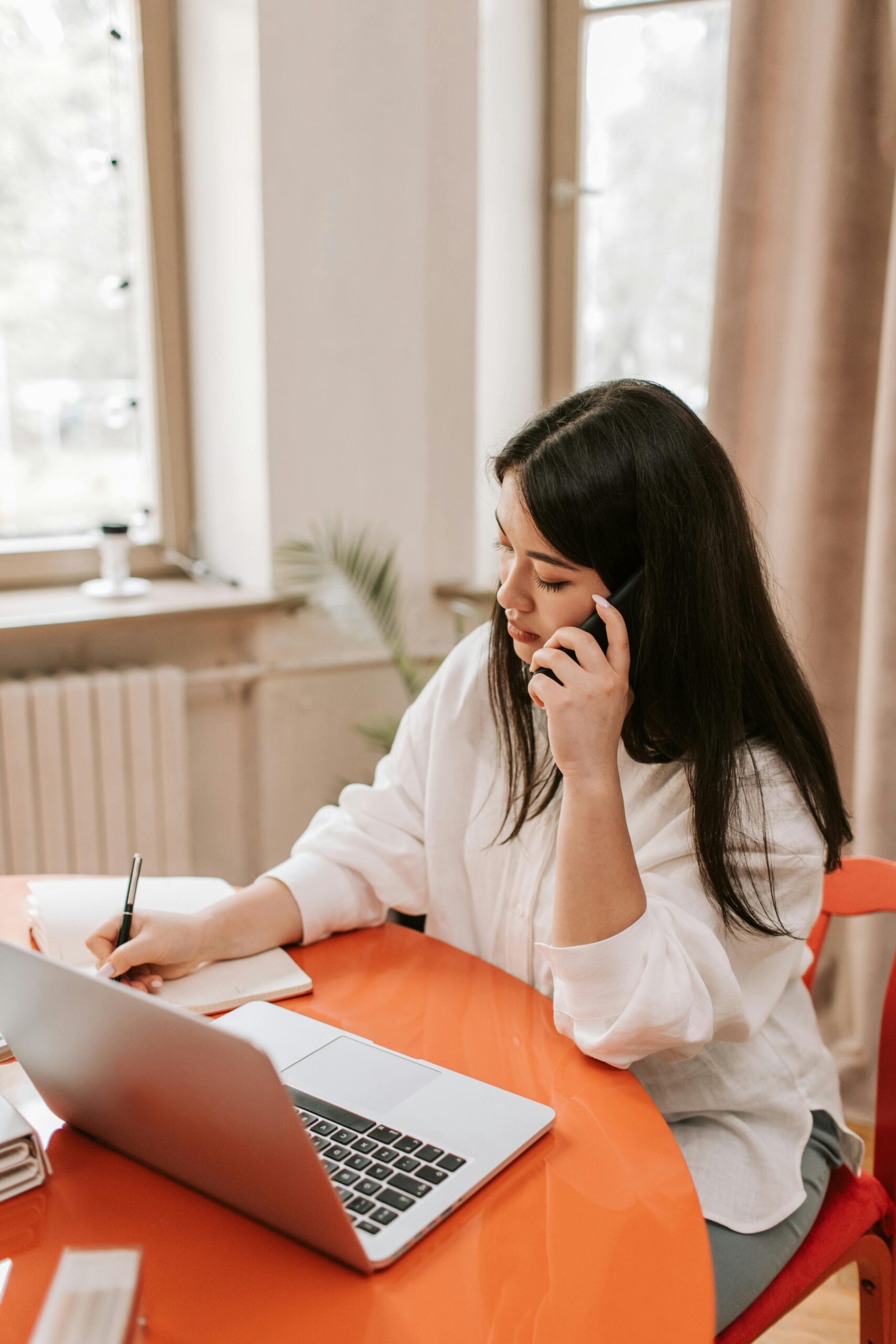 Woman multitasking in a home office, using phone and laptop at a table.