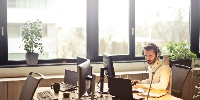 A businessman sits at a desk using multiple computers and a headset in a well-lit modern office.